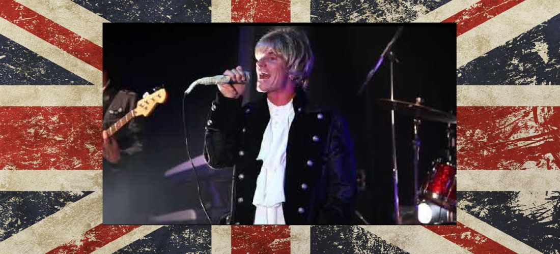 The lead singer of The Who Revue performing live, wearing a classic Roger Daltrey-style outfit. The image is framed against a distressed Union Jack background.
