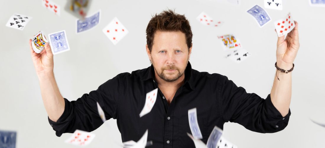 Magician Andy Gross in a black shirt holding playing cards as a deck of cards flies around him.