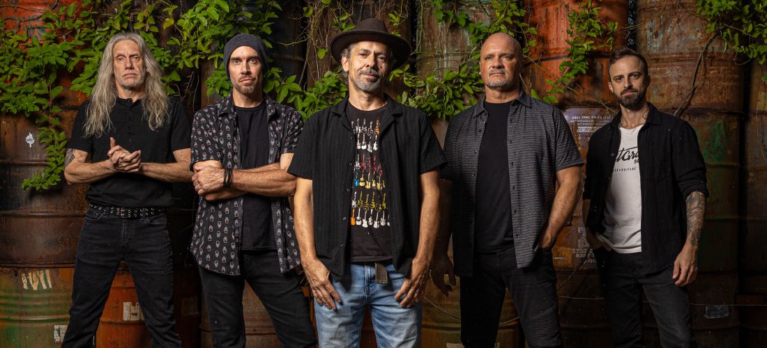 A promotional group portrait of a five-member rock band posing in front of weathered, rusty industrial barrels covered in green vines.