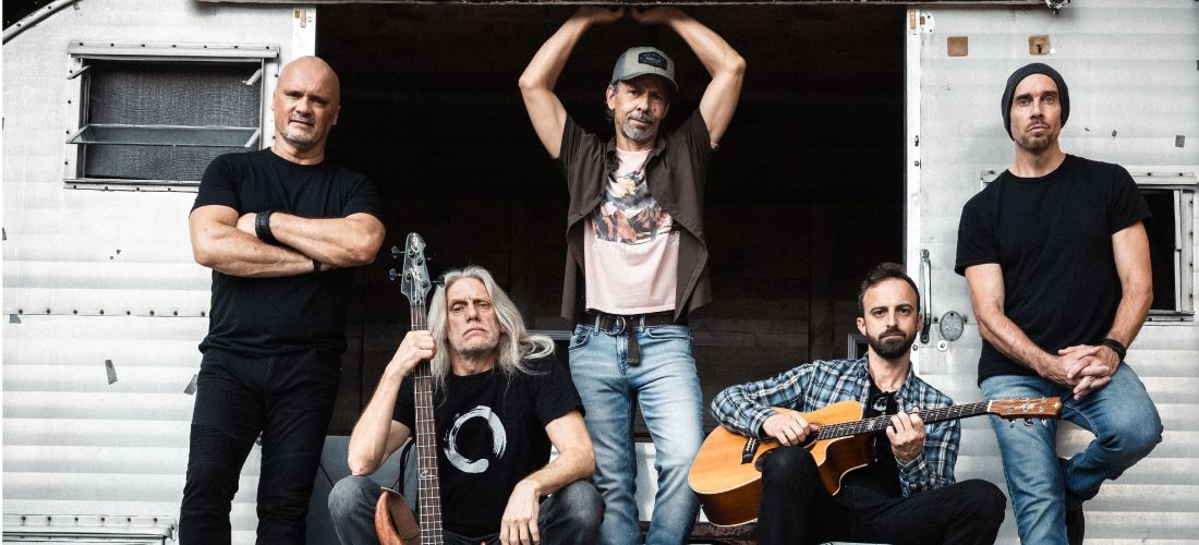 A promotional photo of the five band members posing outdoors in front of a white vintage travel trailer, with two members sitting on the ground holding string instruments.