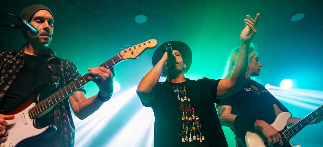 A dynamic, close-up concert photo of three band members performing on a stage illuminated by bright green spotlights and hazy smoke.