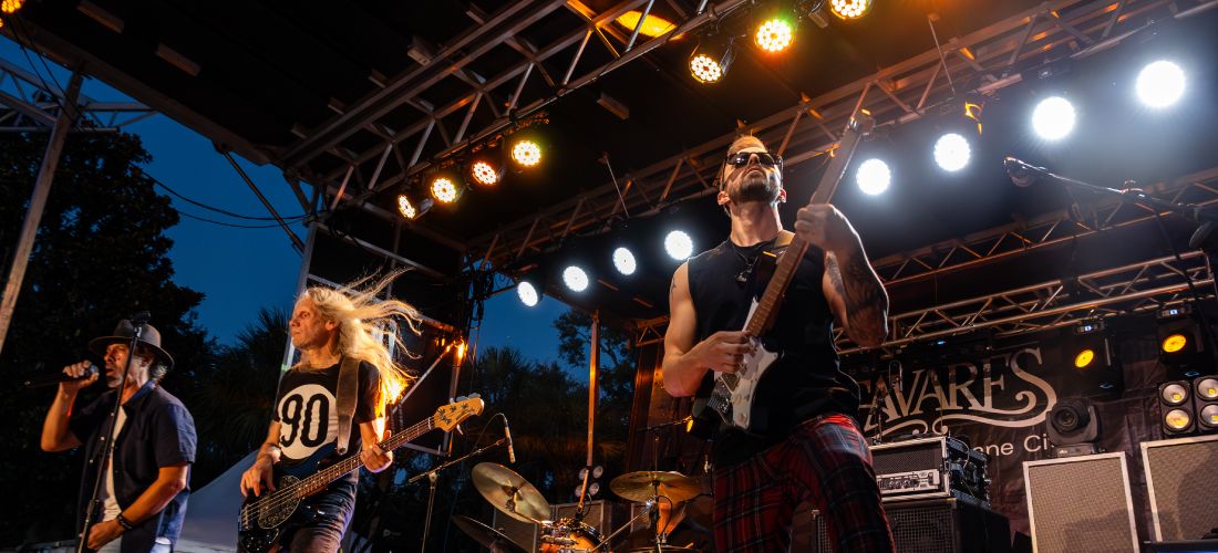 An outdoor evening concert photo capturing the band performing under an elaborate stage lighting rig, with the background banner showing the word "Tavares."