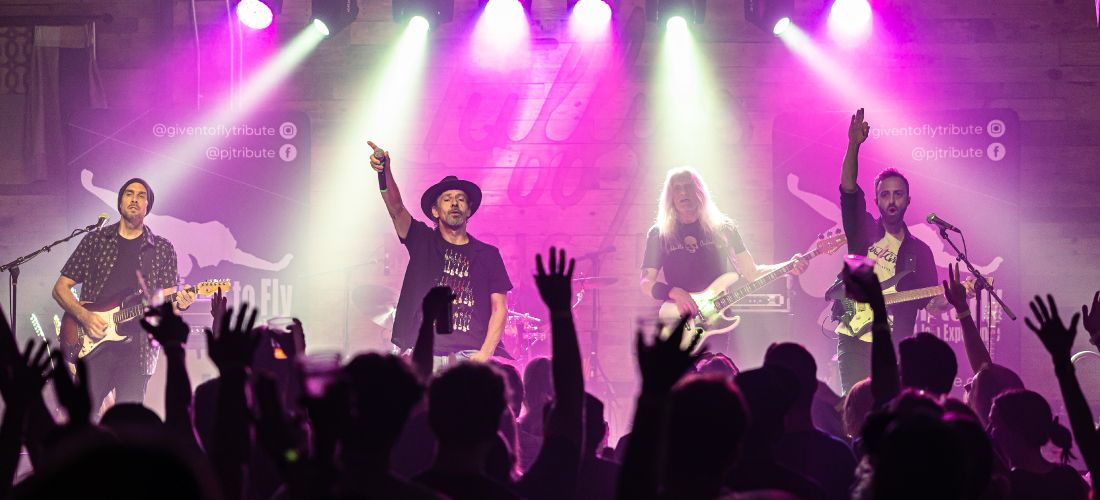 A wide-angle concert photo of the full band playing on a stage under vibrant purple spotlights, viewed from behind a crowd of fans holding their hands in the air.